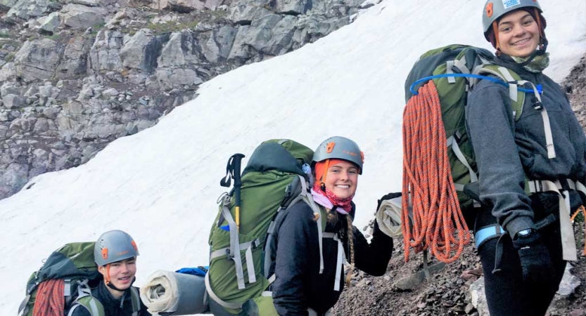 Three people wearing safety gear carry backpacks along a rocky and snowy path